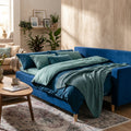 A lifestyle scene illustrating the Noret Sofa bed in a cozy home setting during daytime, shot with a 50mm lens at eye level. The sofa is styled as a bed with plush bedding in complementary oceanic hues, positioned against a textured wall with warm ambient 