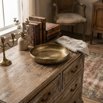Create a lifestyle scene featuring the Ankara Small Round Plate with Brass Finish placed atop a rustic wooden dresser, styled with vintage decor elements for a cozy, homey feel. Use HDR studio lighting to evenly illuminate the plate, capturing its textured