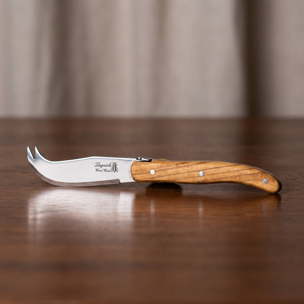 A high-resolution, front-facing studio shot of the Laguiole Wood Short Cheese Knife (HW), delicately placed on a polished dark wood surface with a subtle reflection beneath, showcasing the expertly crafted olive wood handle with intricate grain details. Th