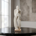 Create a cinematic, high-resolution studio shot of the 'Standing Couple - White' sculpture as a centerpiece on a sleek, reflective black marble table. Use diffused daylight balanced with HDR studio lighting to highlight the smooth, polished wooden texture 