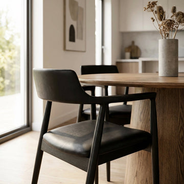 Generate a lifestyle scene featuring the Ealing Dining Chair in Black Leather within a contemporary dining room environment. Depict the chair placed at a stylish wooden dining table, with warm natural daylight streaming through a nearby window, complemente