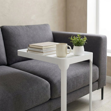 A high-resolution, studio shot of the 'Sofa side table White' positioned next to a plush, modern gray sofa in a contemporary living room setting. The scene is illuminated with softbox lighting to highlight the smooth, matte white finish of the table, empha