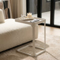 A lifestyle scene inside a chic, well-lit modern living room with warm, inviting tones. The 'Sofa side table White' is styled alongside a cozy cream-colored sofa, situated near large floor-to-ceiling windows that flood the scene with diffused daylight. The