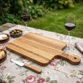 Depict the 'Serving Board Oak' in an outdoor, rustic picnic scene, under natural diffused daylight. Use HDR studio lighting effects to bring out the warm, honey-toned wood grain and the detailed craftsmanship of the board. The shot is a top-down perspectiv