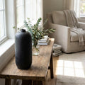A lifestyle scene showcasing the Basington Vase Matte Black placed on a rustic wooden console in a sunlit, cozy living room. Natural diffused daylight filters through large windows, creating soft shadows and highlighting the handcrafted details of the vase