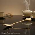 Visualize the Gingko Gold leaf tea spoon artistically arranged on a glossy, reflective surface, shot from a slightly elevated angle with a 50mm lens, capturing the shimmering gold and intricate leaf pattern vividly. Use HDR studio lighting to produce a bal