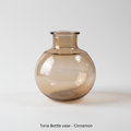 A highly detailed, close-up studio shot of the Turia Bottle Vase - Cinnamon, captured with a macro lens at 100mm focal length. The vase, made from translucent recycled glass with subtle cinnamon and amber hues, is illuminated by softbox lighting to highlig