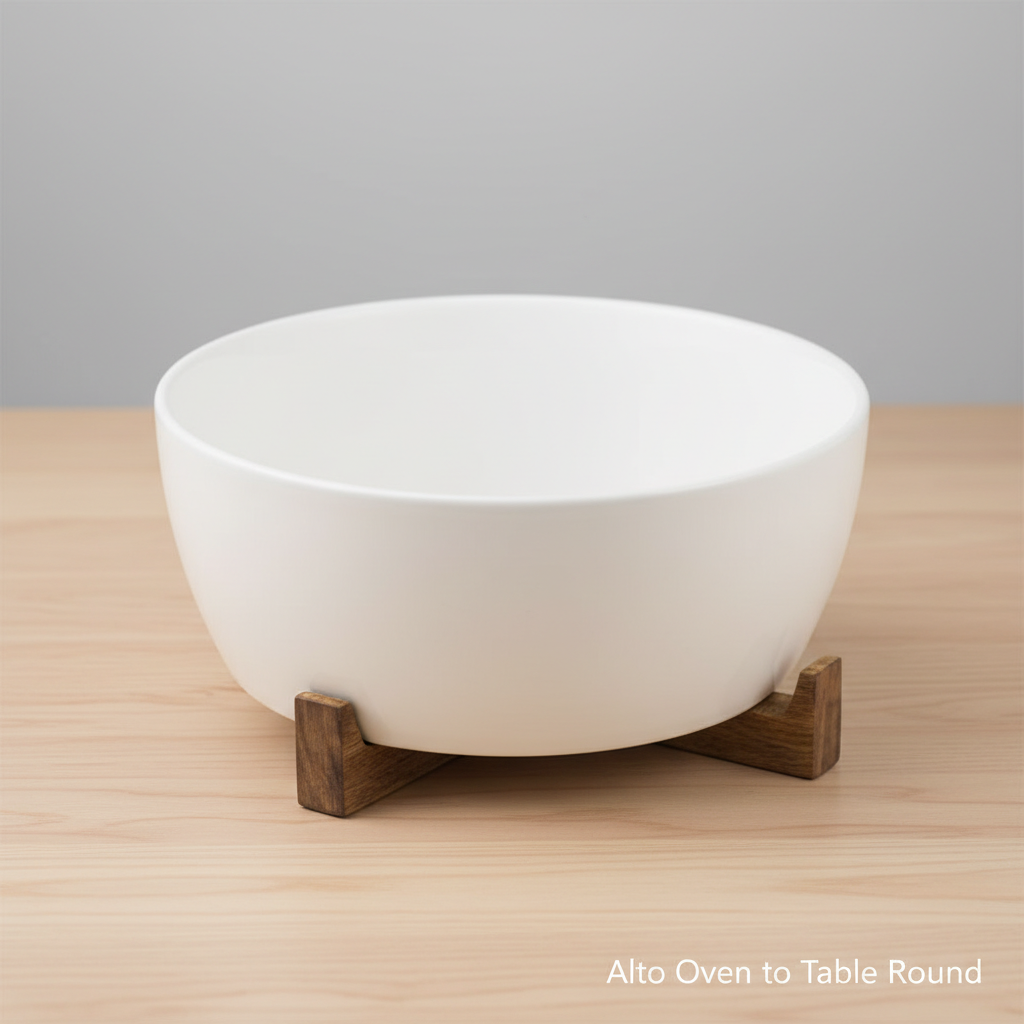 A lifestyle composition featuring the Alto Oven to Table Round dish in a cozy, stylish kitchen setting. Shot with a wide-angle lens at 35mm, capturing the full context of the wooden stand elevating the dish on a well-lit countertop. Natural diffused daylig