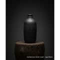 Create a cinematic still of the Earthenware Bottle vessel titled ‘Aged Black’ placed prominently on a textured wooden pedestal in a moody, atmospheric studio setting. Use softbox lighting from above to highlight the vessel’s aged, crackled glaze and uneven