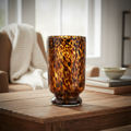 Create a lifestyle scene featuring the 'Tortoise Shell Hurricane. Large' vase, styled on a rustic wooden table in a well-lit, modern living space. Use HDR studio lighting with rim lights to accentuate its amber hues and tortoise shell pattern, creating a l
