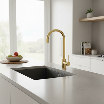A sophisticated lifestyle scene featuring the Mercer SS Gooseneck Mixer in a modern, high-end kitchen environment. Shot with a 24mm wide-angle lens at eye level to capture the overall context, the brass mixer is mounted on a stylish, light-colored quartz c