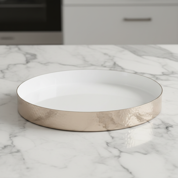 A highly detailed studio shot of the Hammered Round Tray -30cm, White enamel, placed on a sleek marble countertop under softbox lighting that emphasizes its glossy white finish and hammered texture. The camera uses a macro lens at 50mm, with a shallow dept
