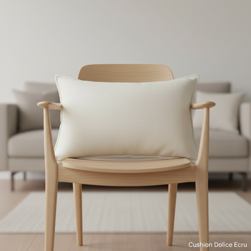 Capture a sophisticated studio shot of the 'Cushion Dolice Ecru', featuring softbox lighting to highlight its plush texture and high-quality fabric. The cushion is centered on a sleek, minimalist wooden chair in a modern living room setting, with a blurred