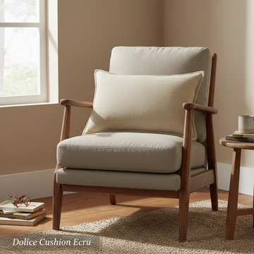 Create a stylized, lifestyle shot of the Dolice Cushion Ecru placed on a rustic, vintage-inspired armchair in a sunlit room. Use diffused daylight to soften the scene, emphasizing the cushion’s handwoven texture and slightly washed-out stonewash look. Posi