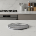 Create a high-resolution, studio-lit image of a 'Marble Hot Plate - Grey' positioned on a sleek, modern kitchen countertop. Use a softbox lighting setup to highlight the smooth, polished marble surface with subtle veining details, emphasizing the cool grey