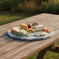 Create a lifestyle shot of the 'Marble Hot Plate - Grey' outdoors on a rustic wooden table, bathed in diffused daylight for a warm, inviting ambiance. Position the hot plate with an assortment of cheeses and small appetizers on it, illustrating its multifu