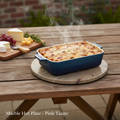 A lifestyle scene featuring the Marble Hot Plate - Pink Taupe, styled on a rustic outdoor wooden table under diffused daylight that enhances the warm pink taupe tones and marble veining. The hot plate is placed beneath a baking dish with steaming contents,